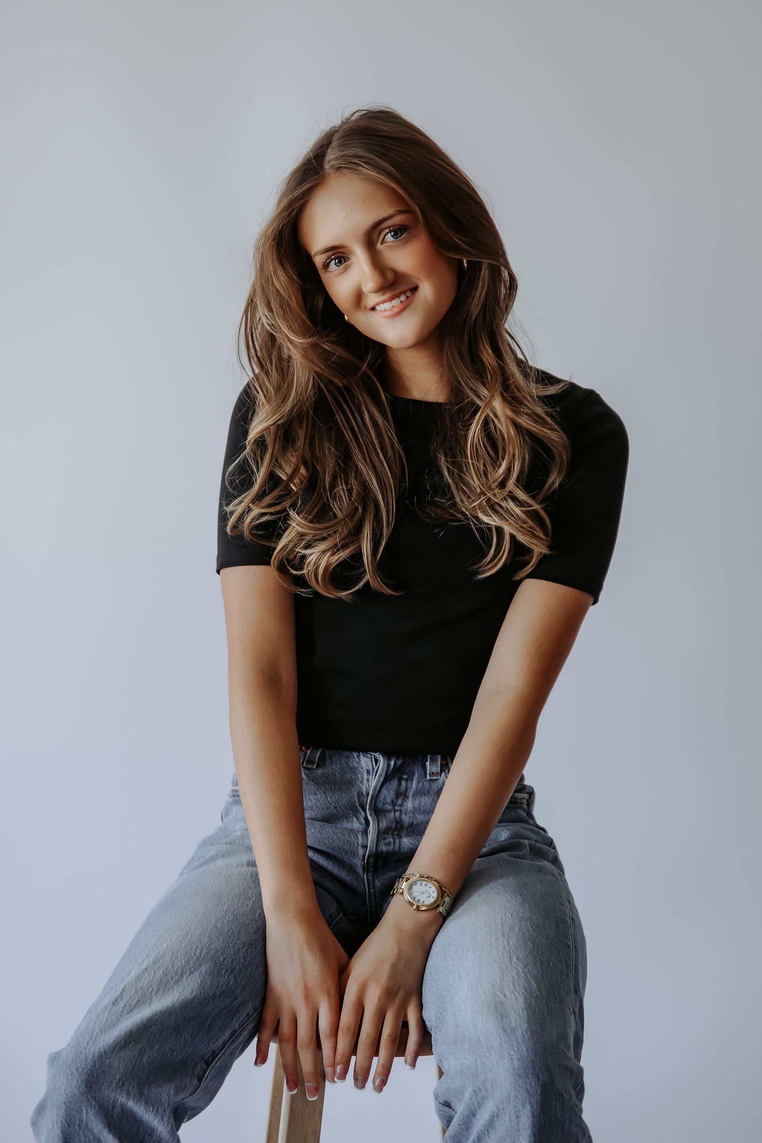 Smiling young woman with wavy brown hair wearing black top and jeans sitting on stool with watch on wrist.