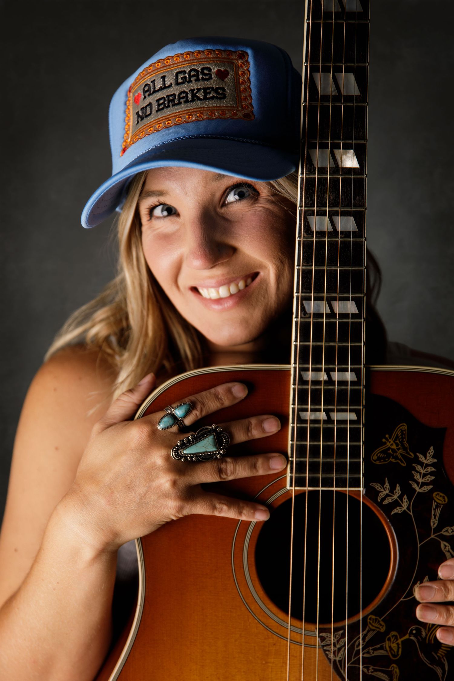 Smiling woman holding acoustic guitar, wearing blue trucker hat reading All Gas No Brakes, with turquoise rings.