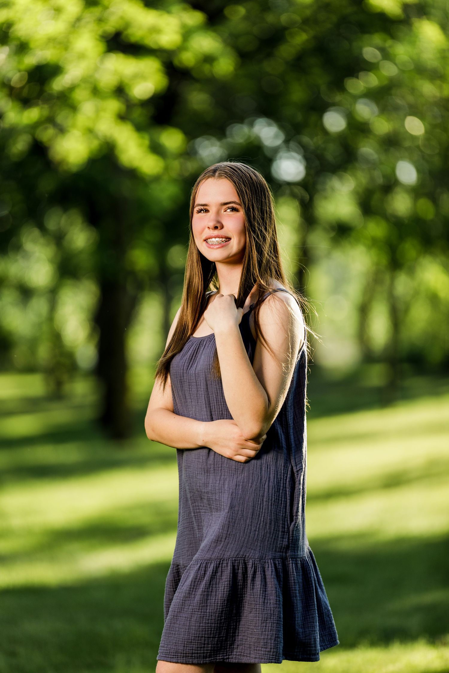 Teenage girl in a gray dress posing confidently in a bright green park with bokeh background.