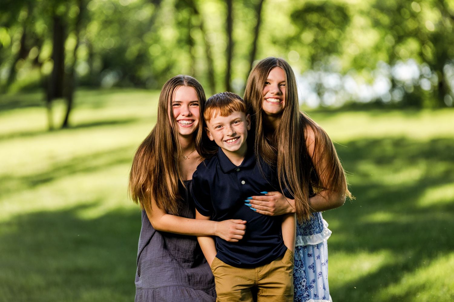 Two teenage girls with long brown hair hug a smiling redhead boy in a sunny green park.