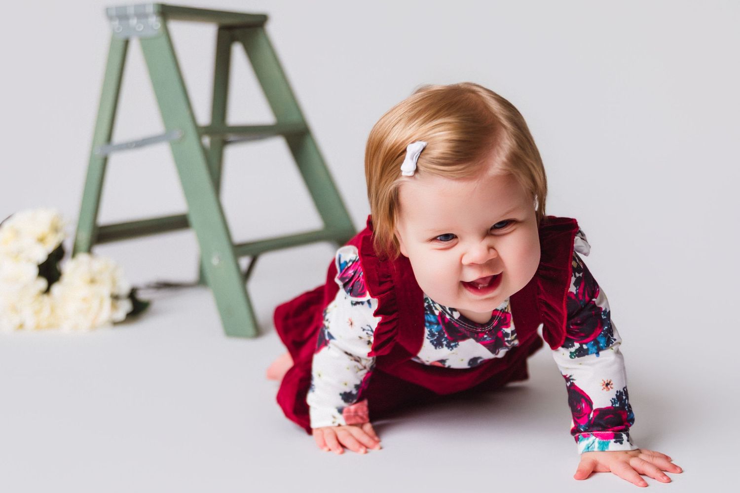 Baby in red floral outfit crawling near a green ladder with flowers.