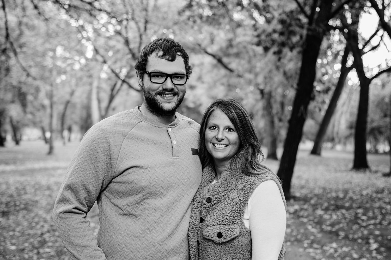A couple poses together for a black and white portrait in a park with fall foliage in the background.