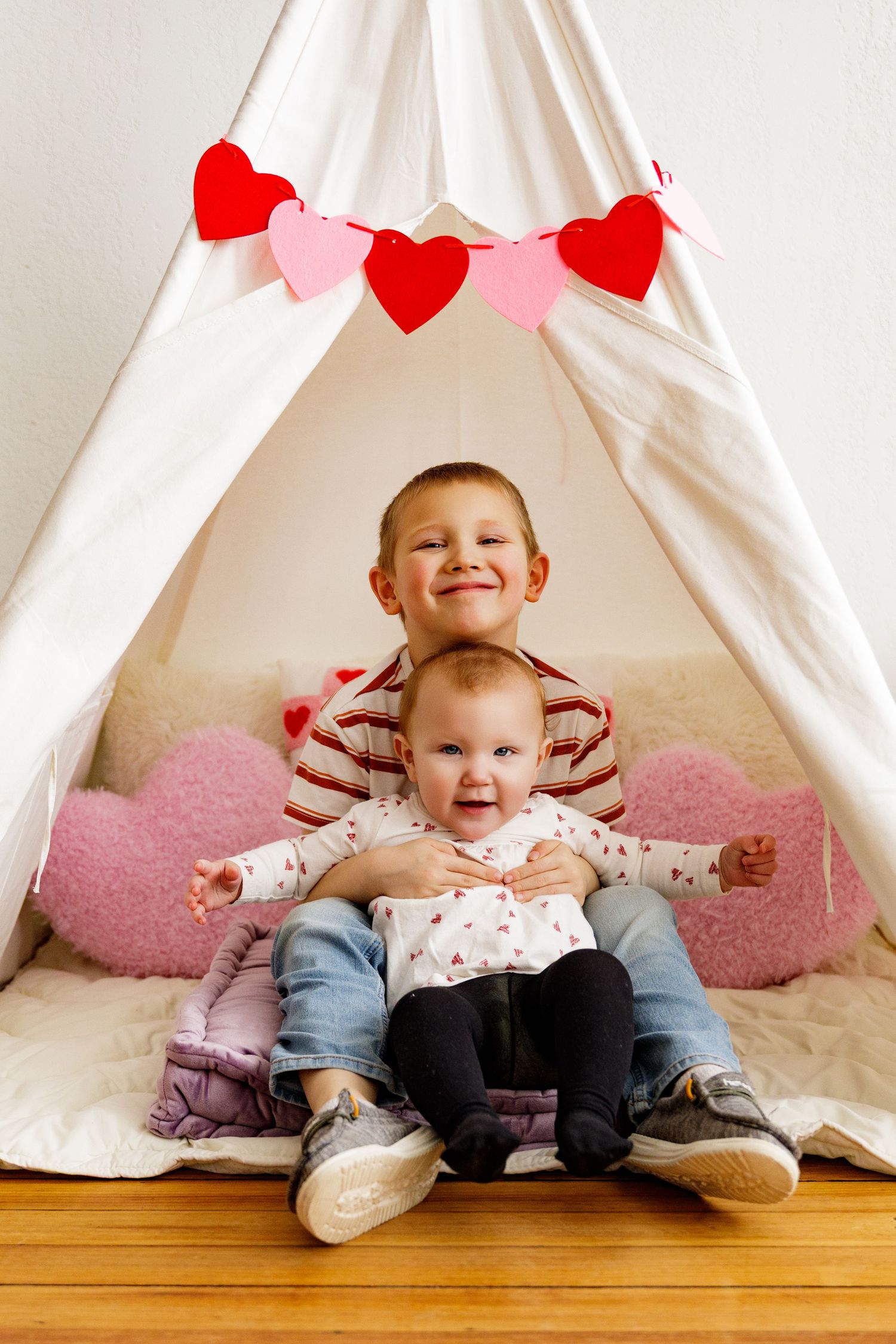 Valentine's themed photo series with a baby sitting under a white teepee decorated with red heart garland.