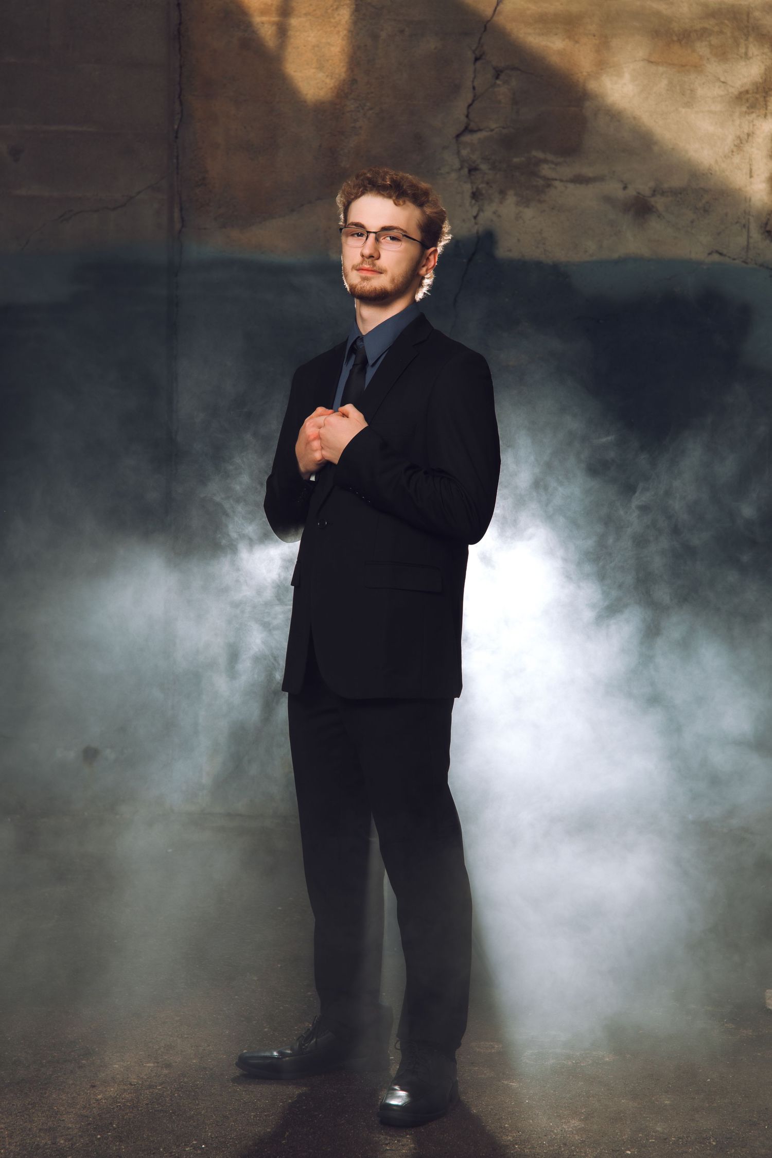 Young man in black suit and glasses poses confidently in smoky dramatic lighting against a textured wall.