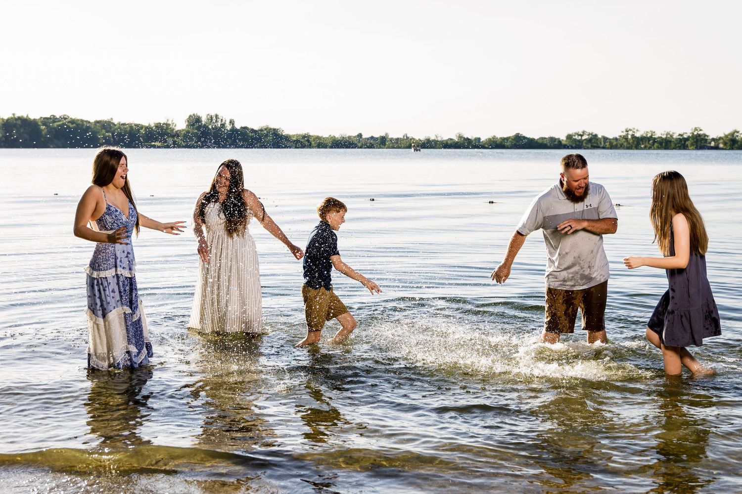 Joyful family holding hands and wading through shallow lake water during a summer photo shoot.