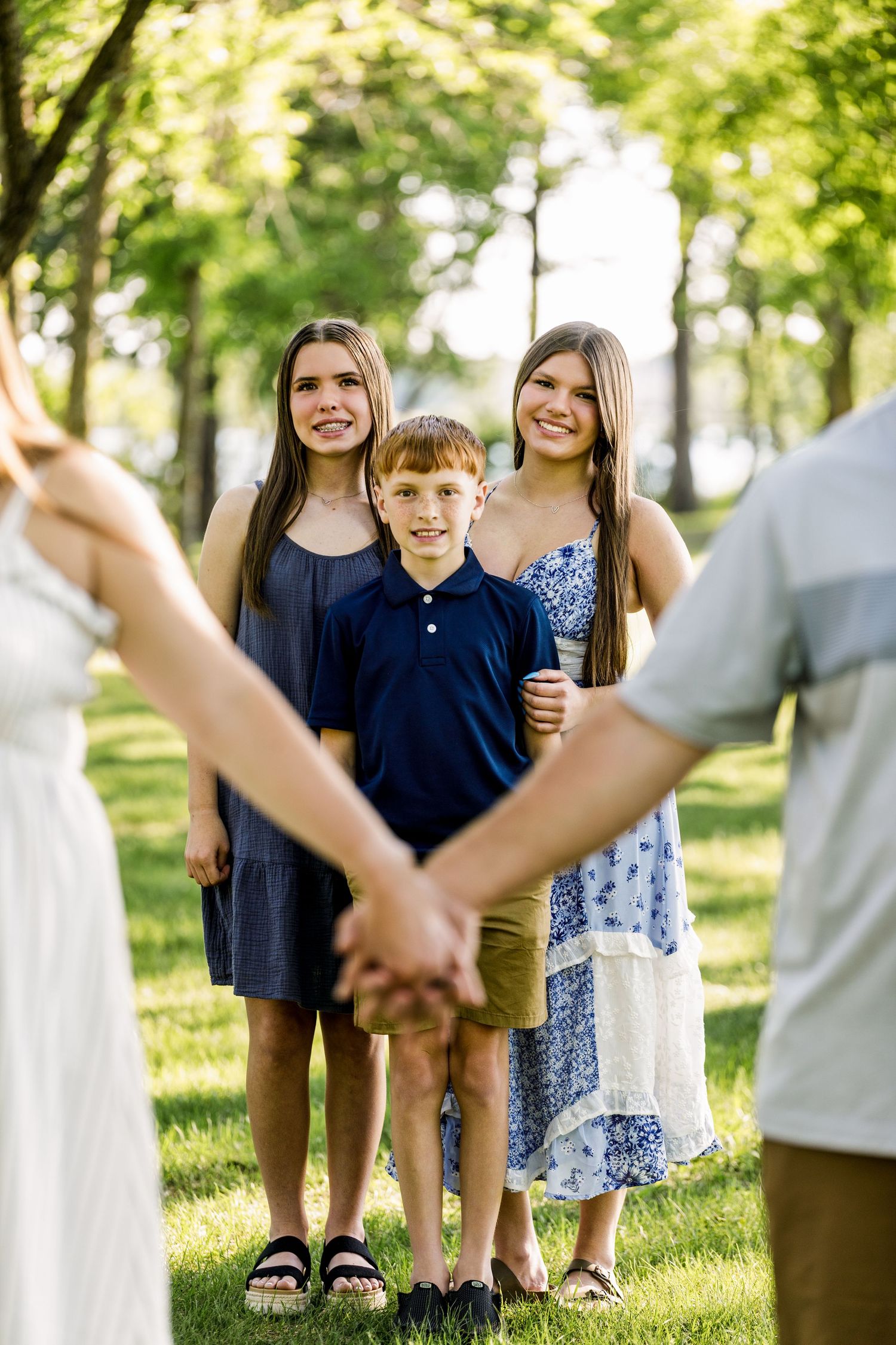 Family of five smiling in a sunny park, two adults holding hands in foreground with three children behind them.