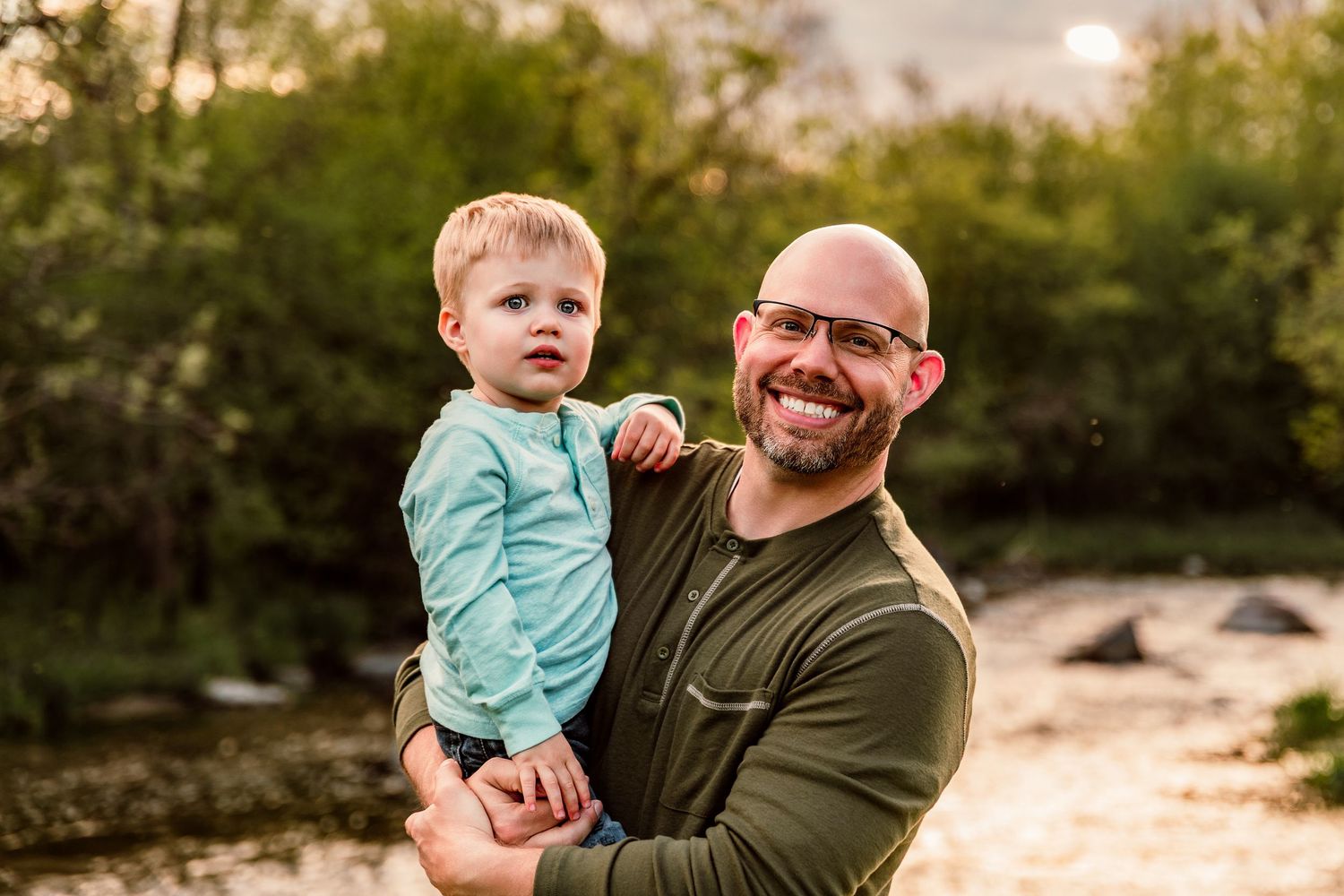 Smiling bald man with glasses holds a toddler in a teal shirt outdoors near a creek at golden hour.