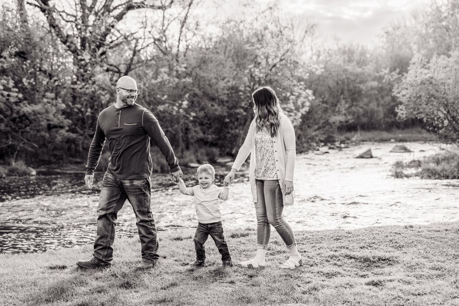 Black and white photo of a family with a toddler walking outdoors near a creek, holding hands in a natural setting.