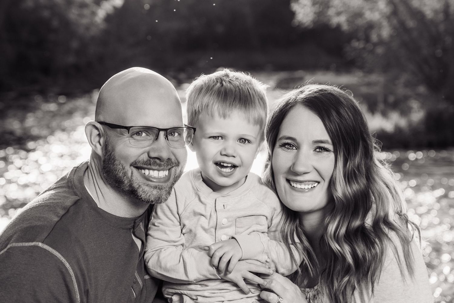 Black and white family portrait of smiling parents with young toddler boy outdoors near a sunlit river.