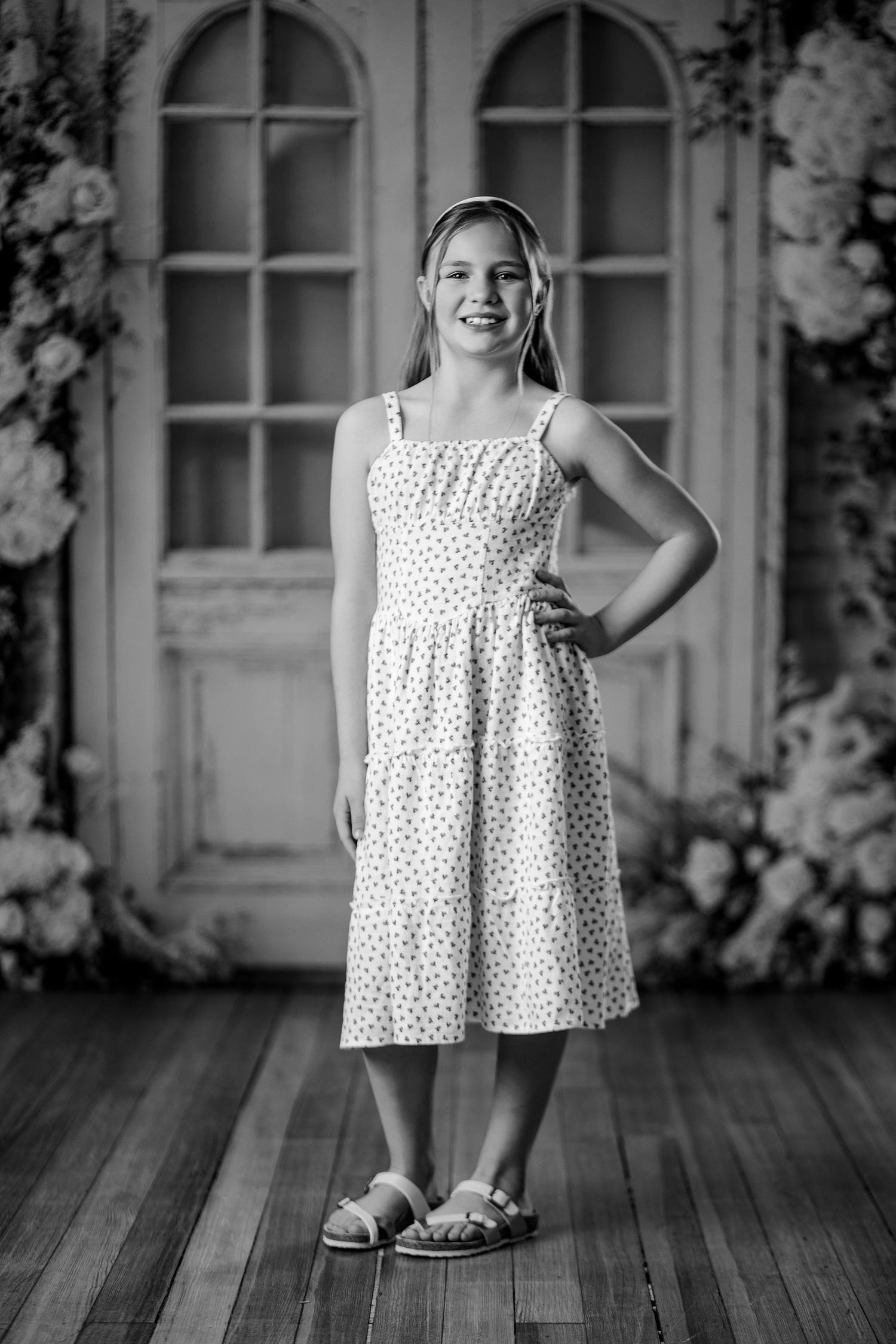 Young girl in polka dot sundress poses confidently in black and white portrait with floral backdrop.