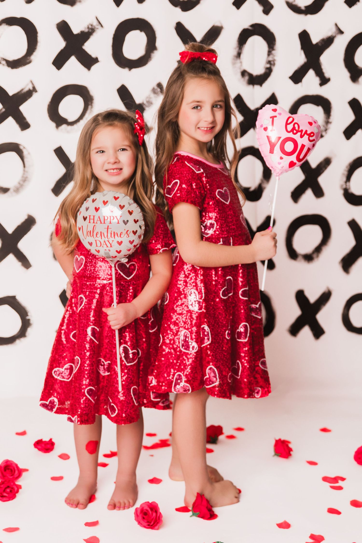 Two girls in matching red sequin heart dresses hold Valentine's Day balloons against an XO patterned backdrop.