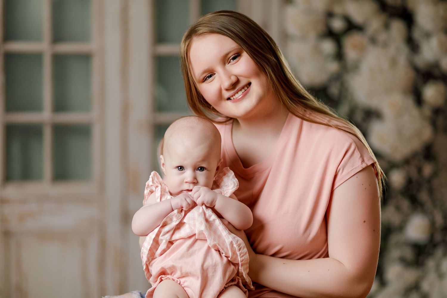 A smiling young woman in a pink top holds a cute baby girl in a floral dress during a professional photo session.