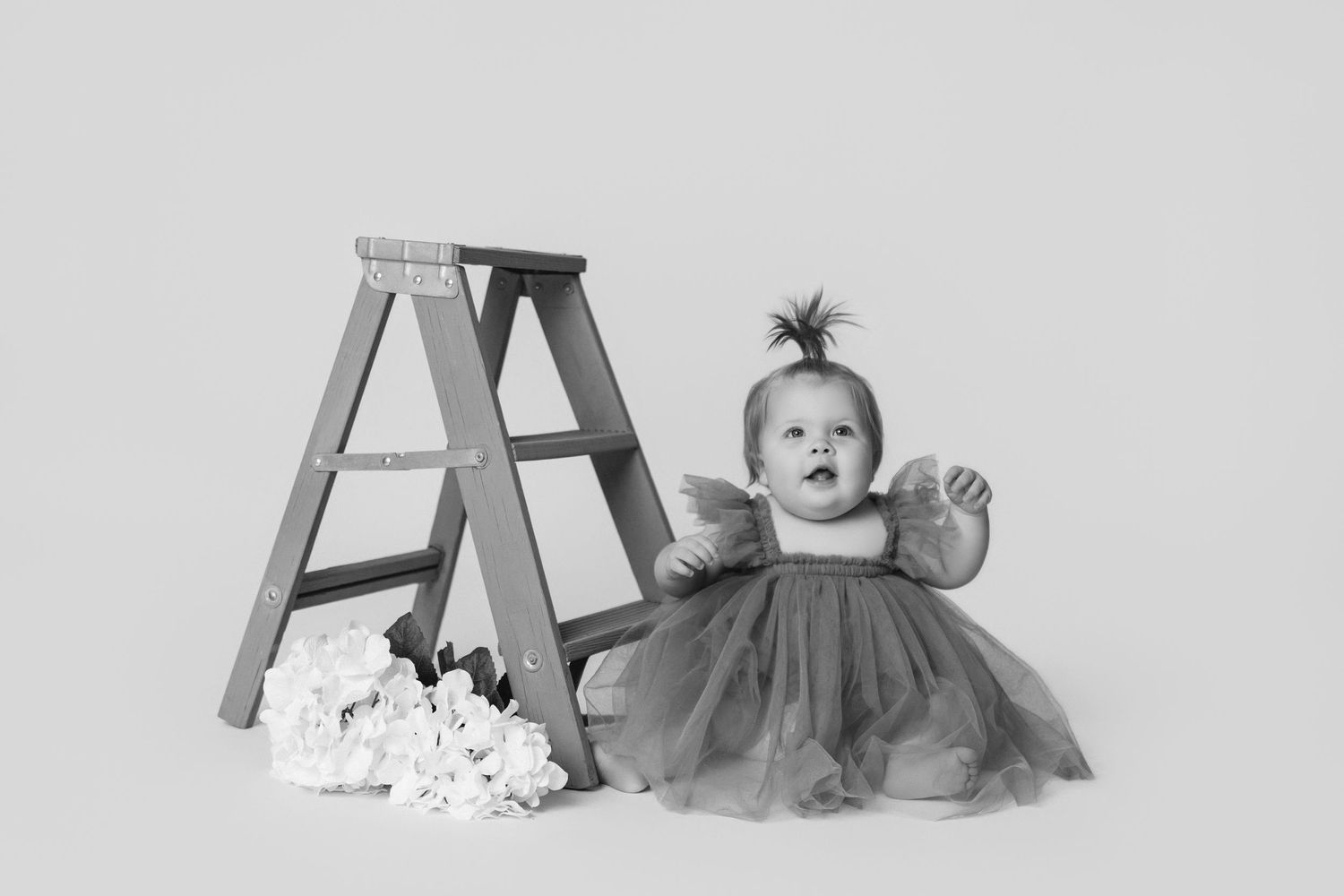 Baby in tulle dress exploring near wooden ladder prop in studio.