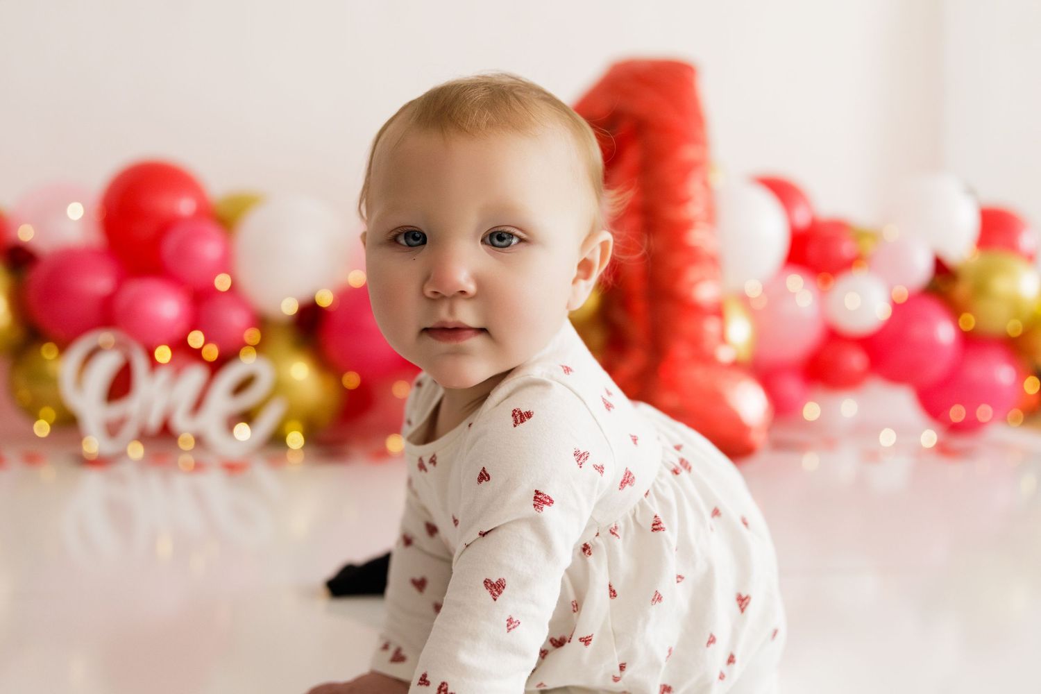 Baby in white outfit sits in front of festive red and gold balloon display with number one decoration.