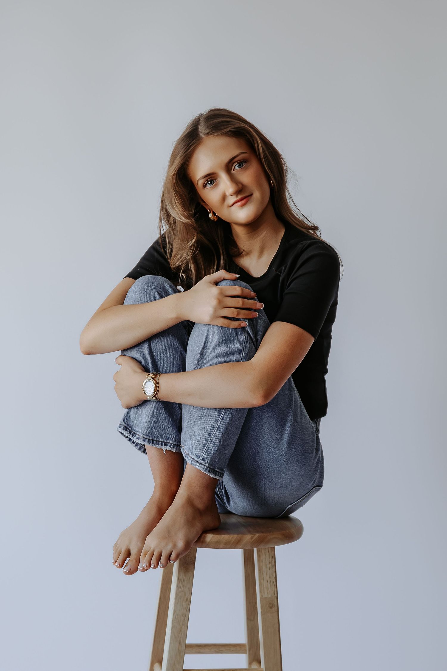 Young woman sitting on wooden stool hugging knees, wearing black tee and jeans, barefoot against gray background.