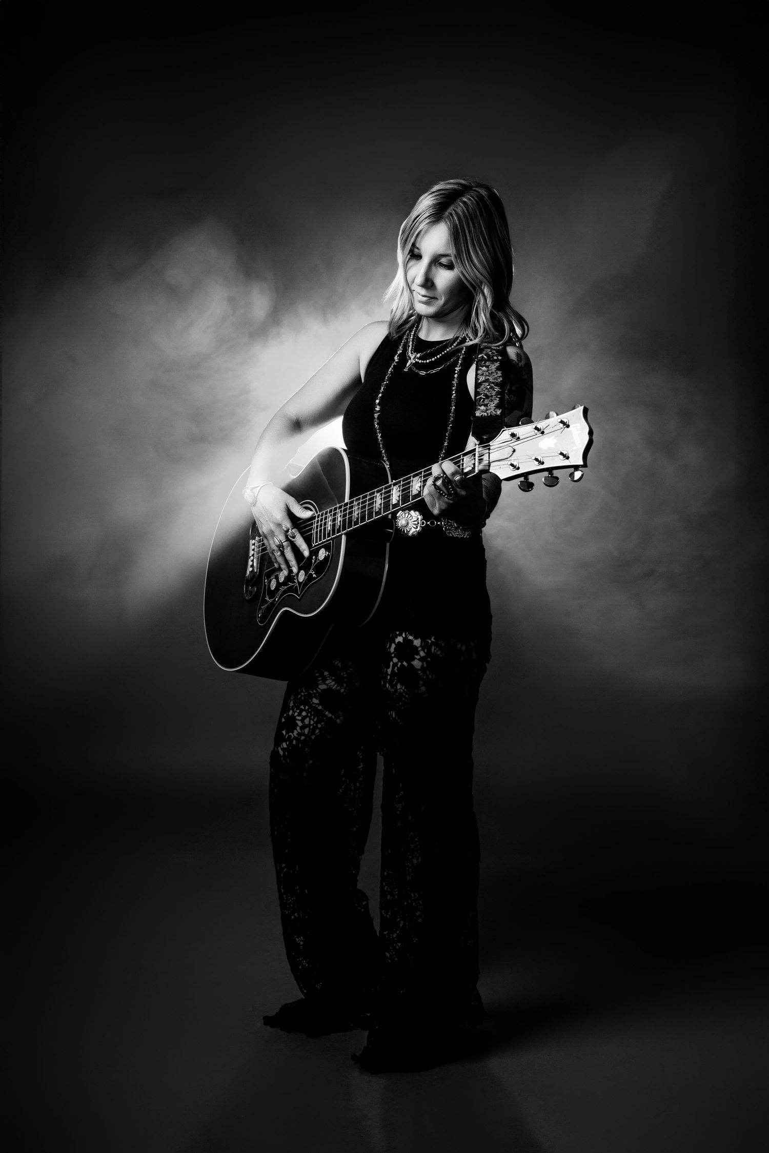 Black and white photo of a tattooed blonde woman playing an acoustic guitar while standing in a dramatic smoky light.