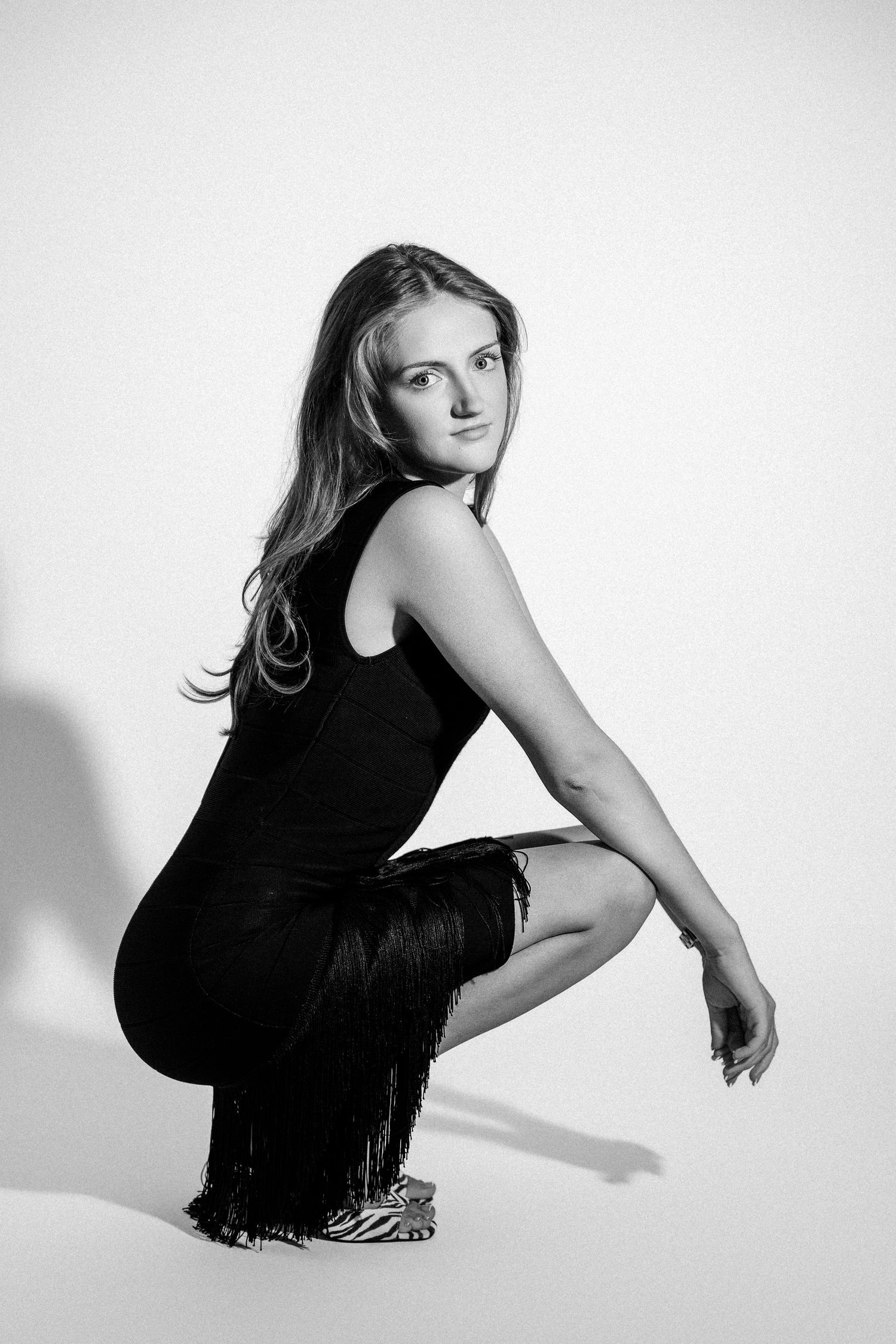 Black and white photo of a young woman crouching in a black fringe dress and zebra print heels.