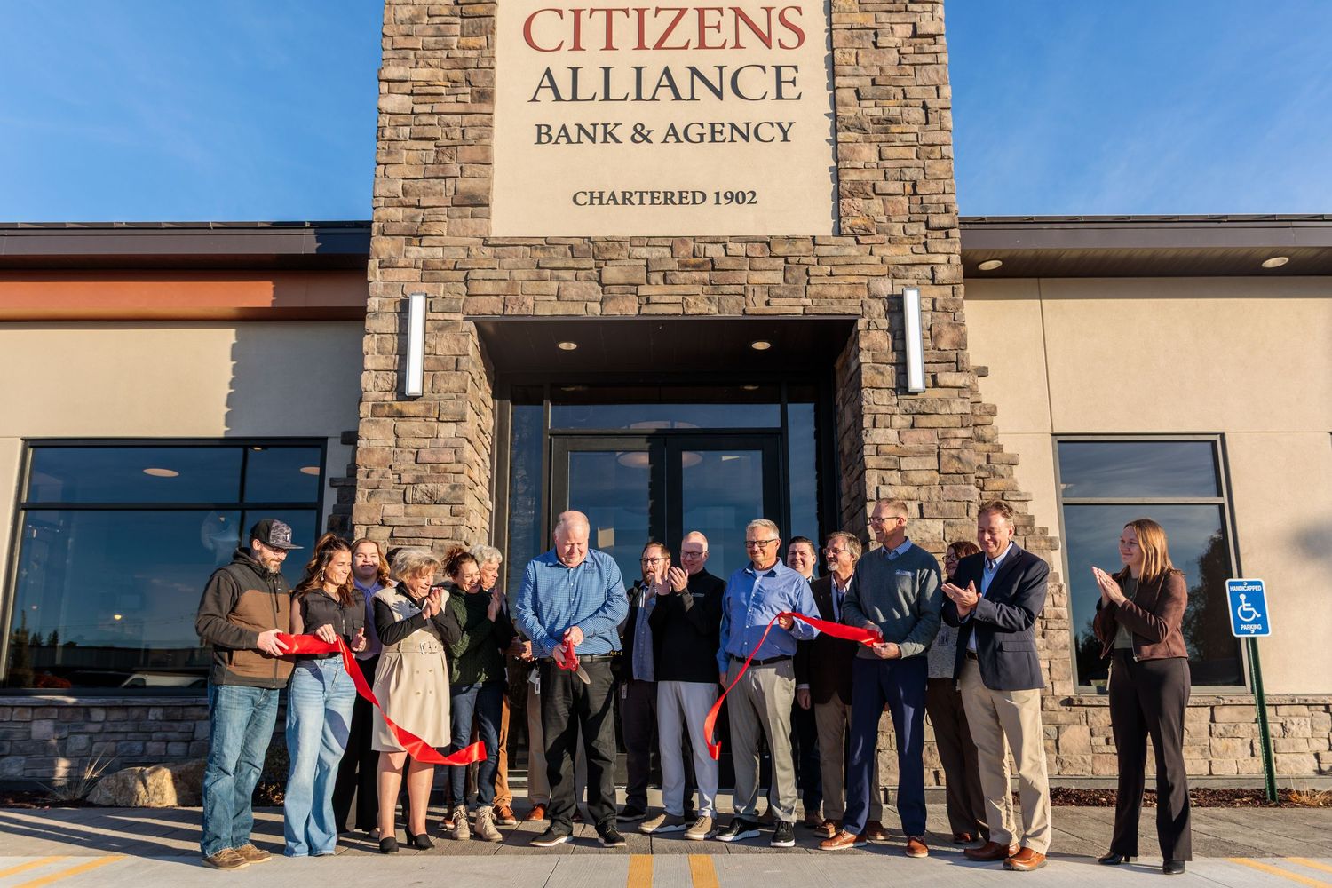 Group of people celebrating a ribbon cutting ceremony outside Citizens Alliance Bank & Agency, chartered 1902.
