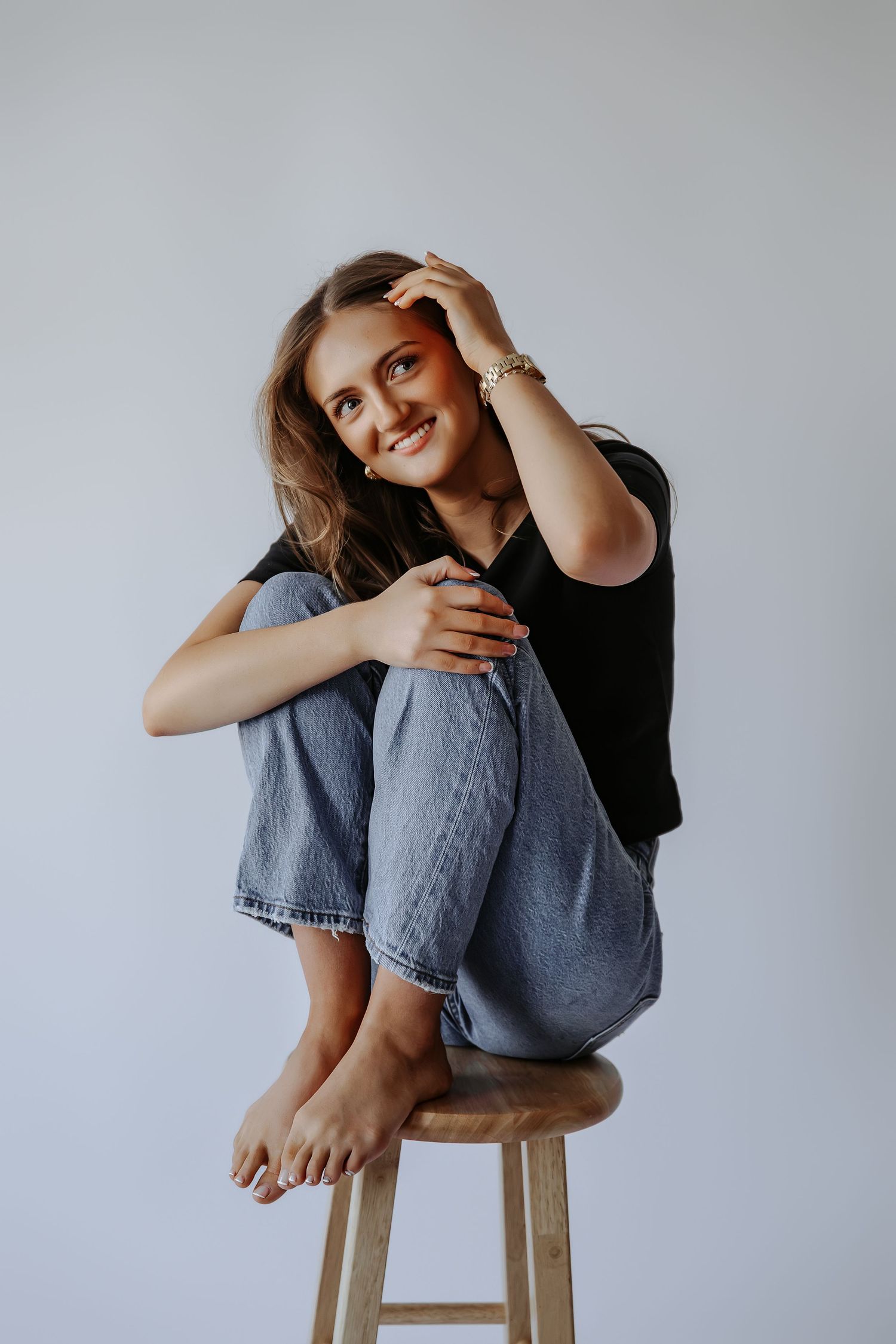 Smiling young woman seated on stool in black top and jeans, hand in hair, colorful eye makeup, barefoot on gray background.