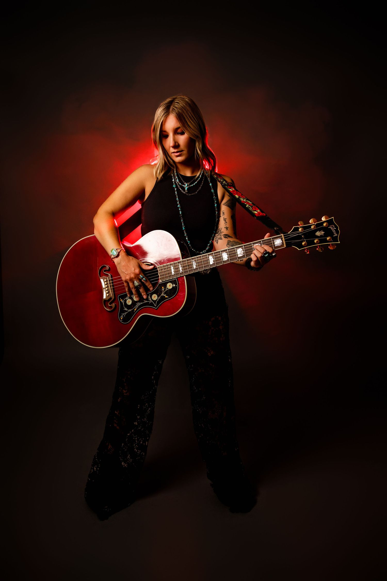 Female guitarist in black outfit plays red acoustic guitar against dramatic dark background with red lighting.