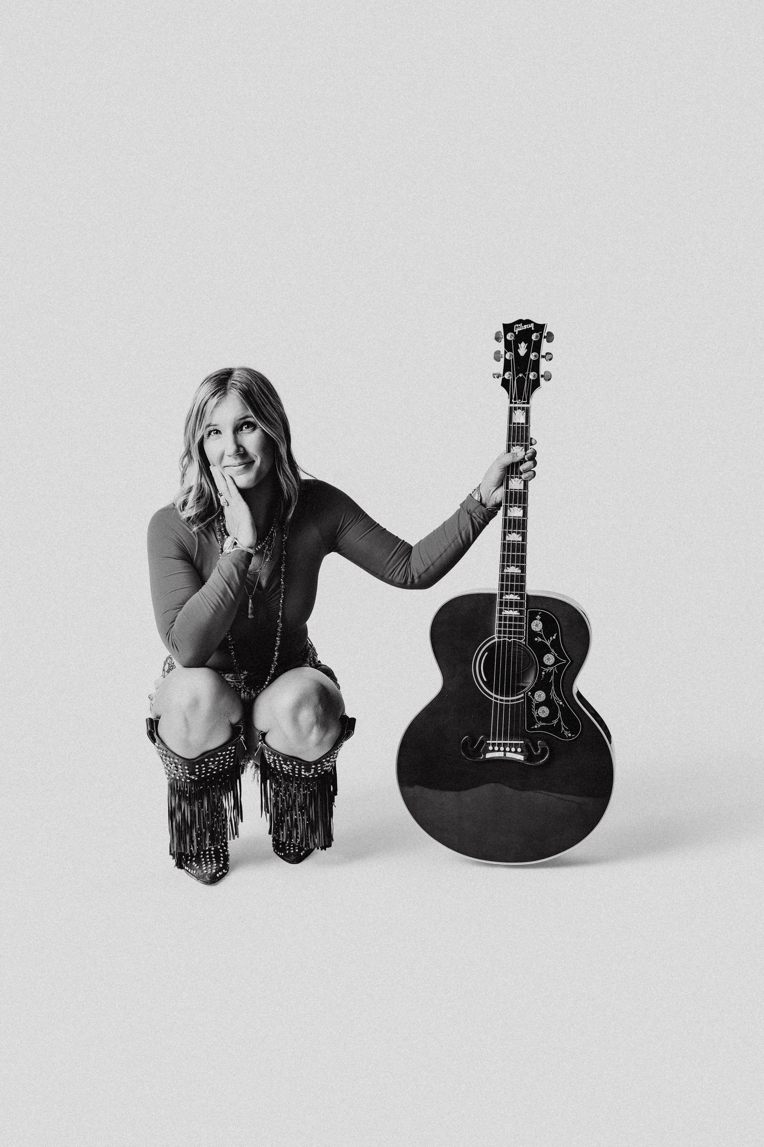 Woman crouching beside a black acoustic guitar in a black and white studio photo wearing fringe boots.