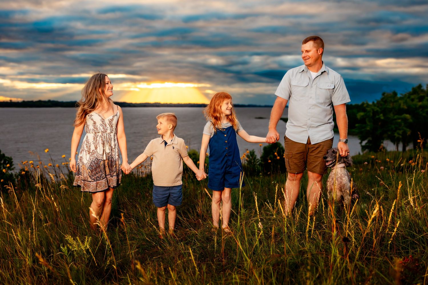 A family holds hands while walking through tall grass at sunset by a waterfront with dramatic clouds overhead.