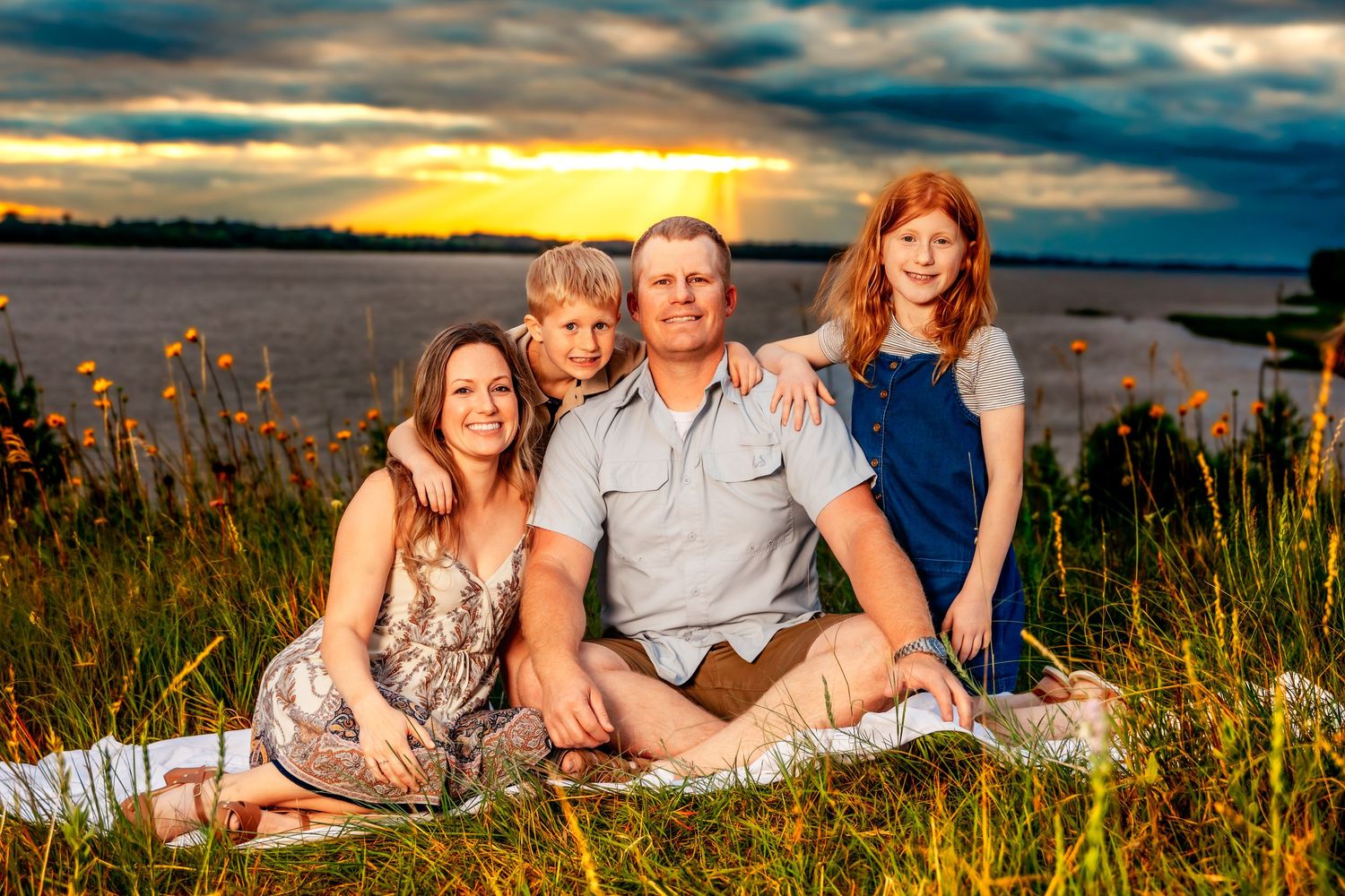 A group gathers on a blanket by the lake at sunset with tall grasses and dramatic clouds in the background.
