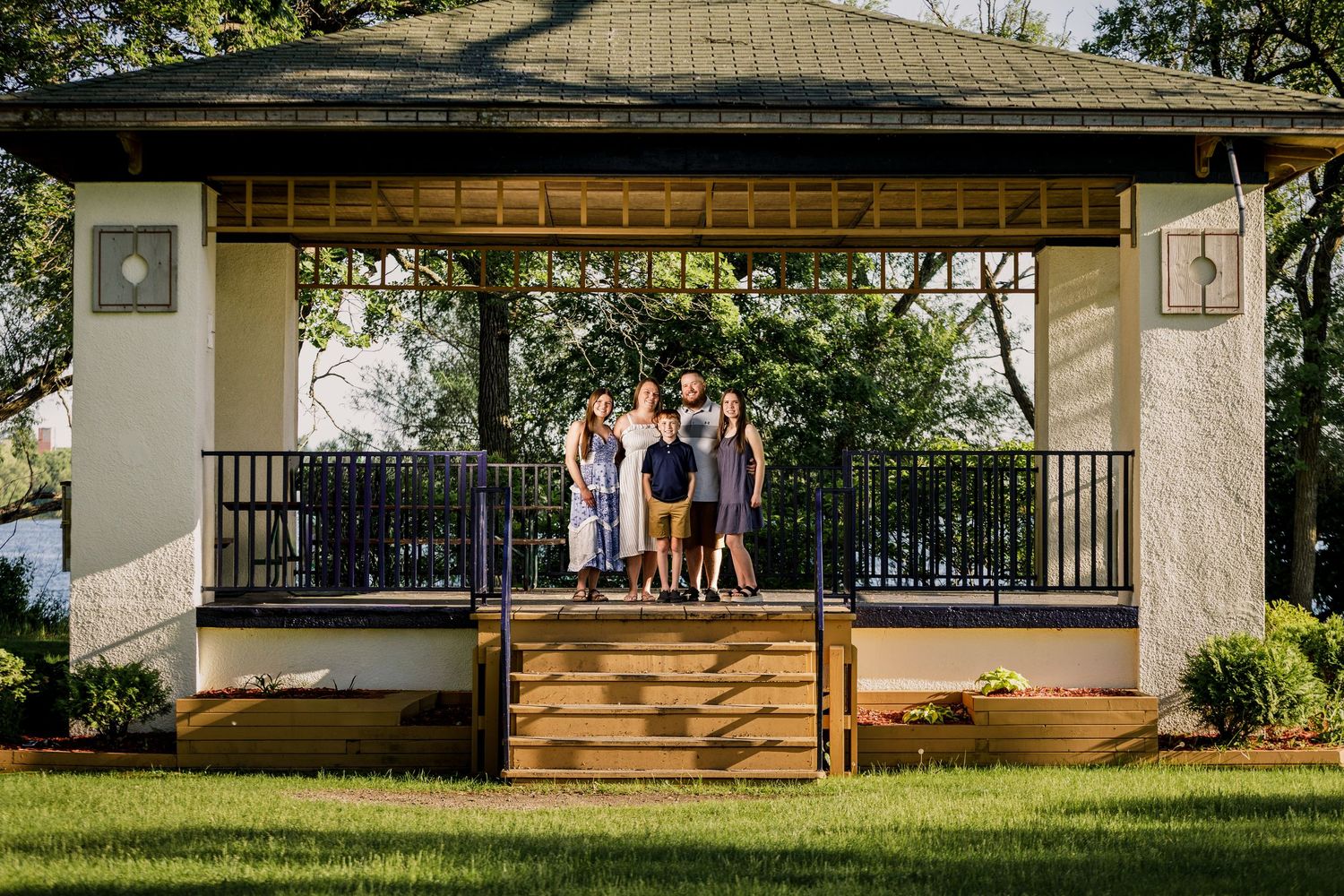 Family of five posing together under a wooden gazebo with black railings in a lush green backyard.