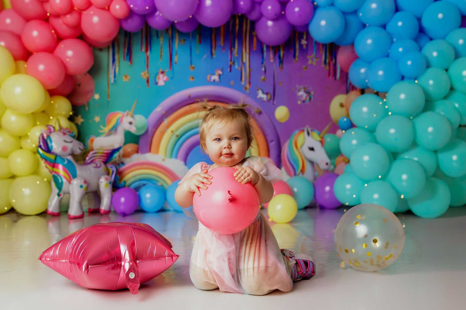 Adorable baby girl sits among rainbow balloon arch and unicorn decorations holding a pink balloon at birthday party.