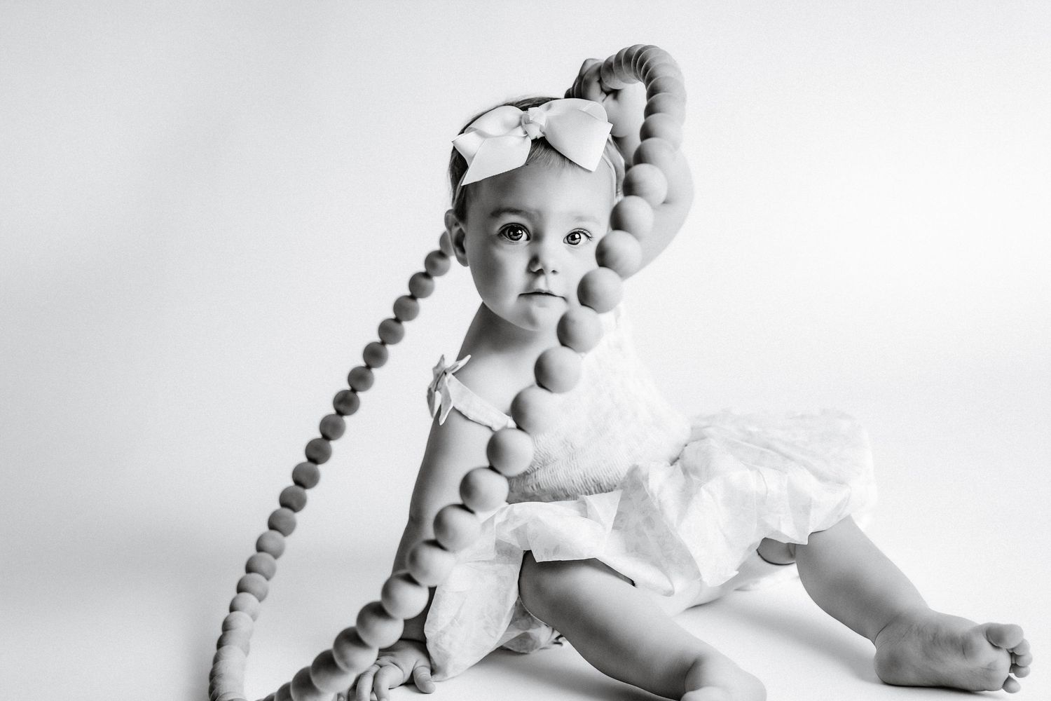 Black and white photo of a cute baby girl in white dress with oversized decorative beaded necklace and bow headband.