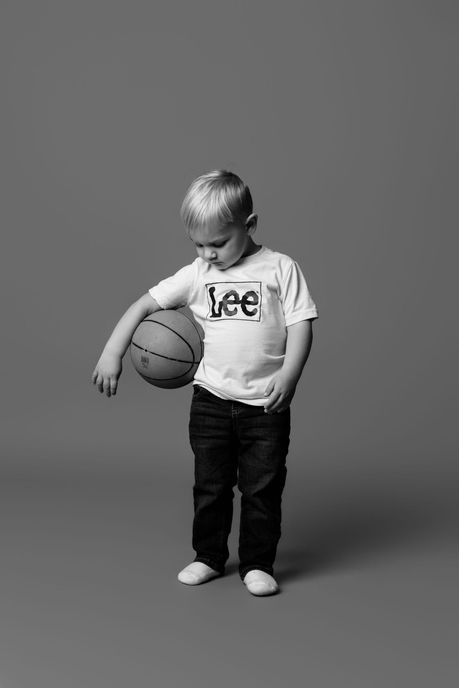 Young child in Lee t-shirt and jeans looking down at basketball against gray studio background.