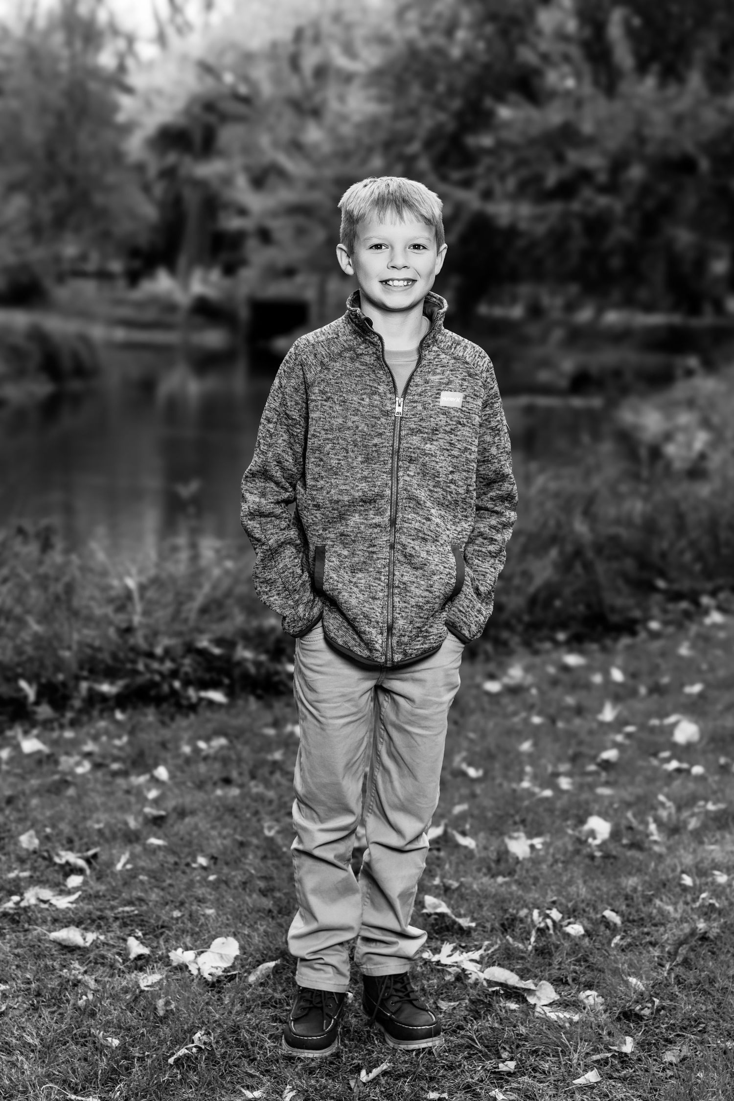 Young child wearing a gray jacket and khaki pants stands smiling on fallen autumn leaves in a park setting.