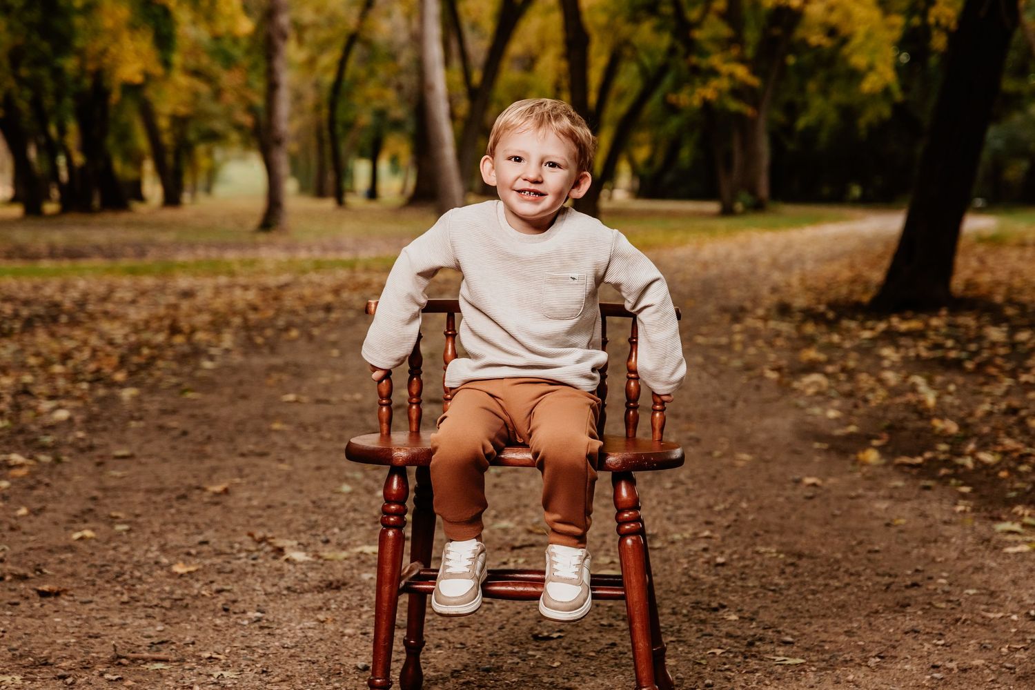 Young child poses happily in brown wooden chair during fall photoshoot.