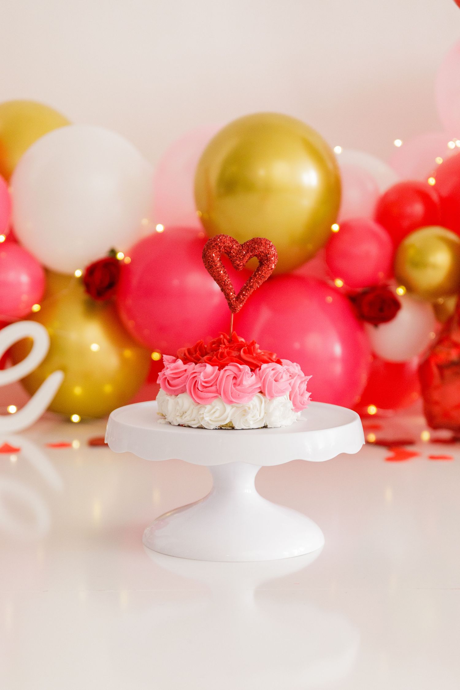 Elegant white cake stand with Valentine's decorations and pink and gold balloons in the background.