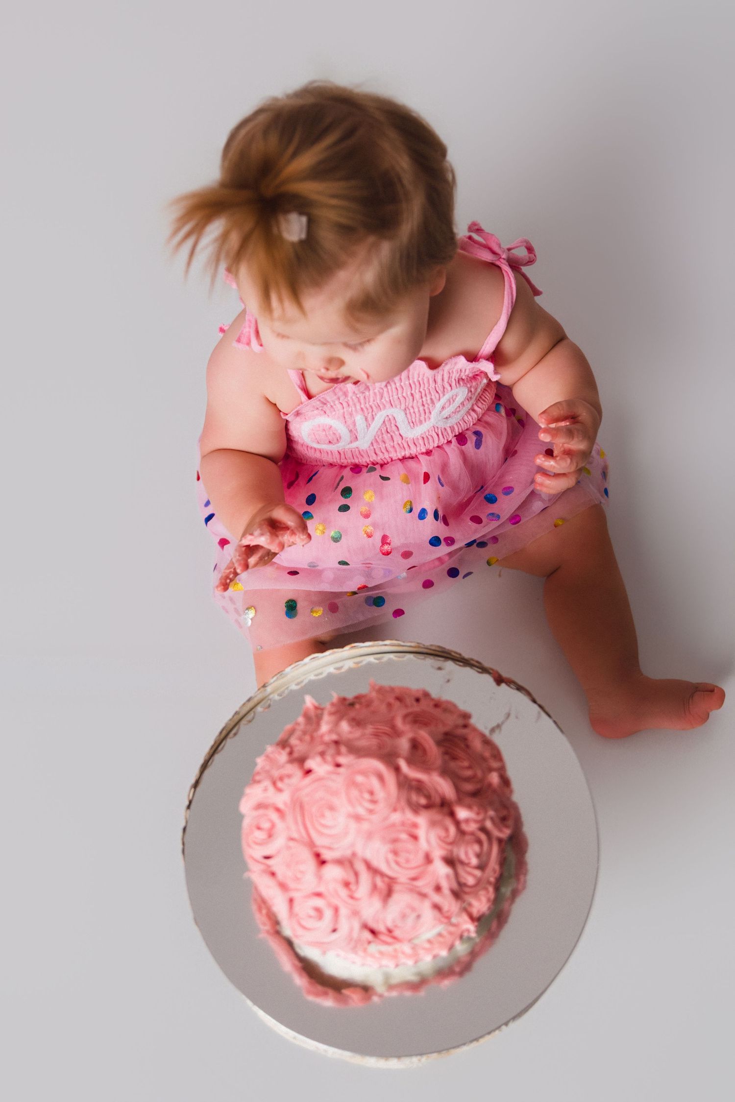 A baby in a pink polka dot dress looks down at a pink frosted cake on a white background.