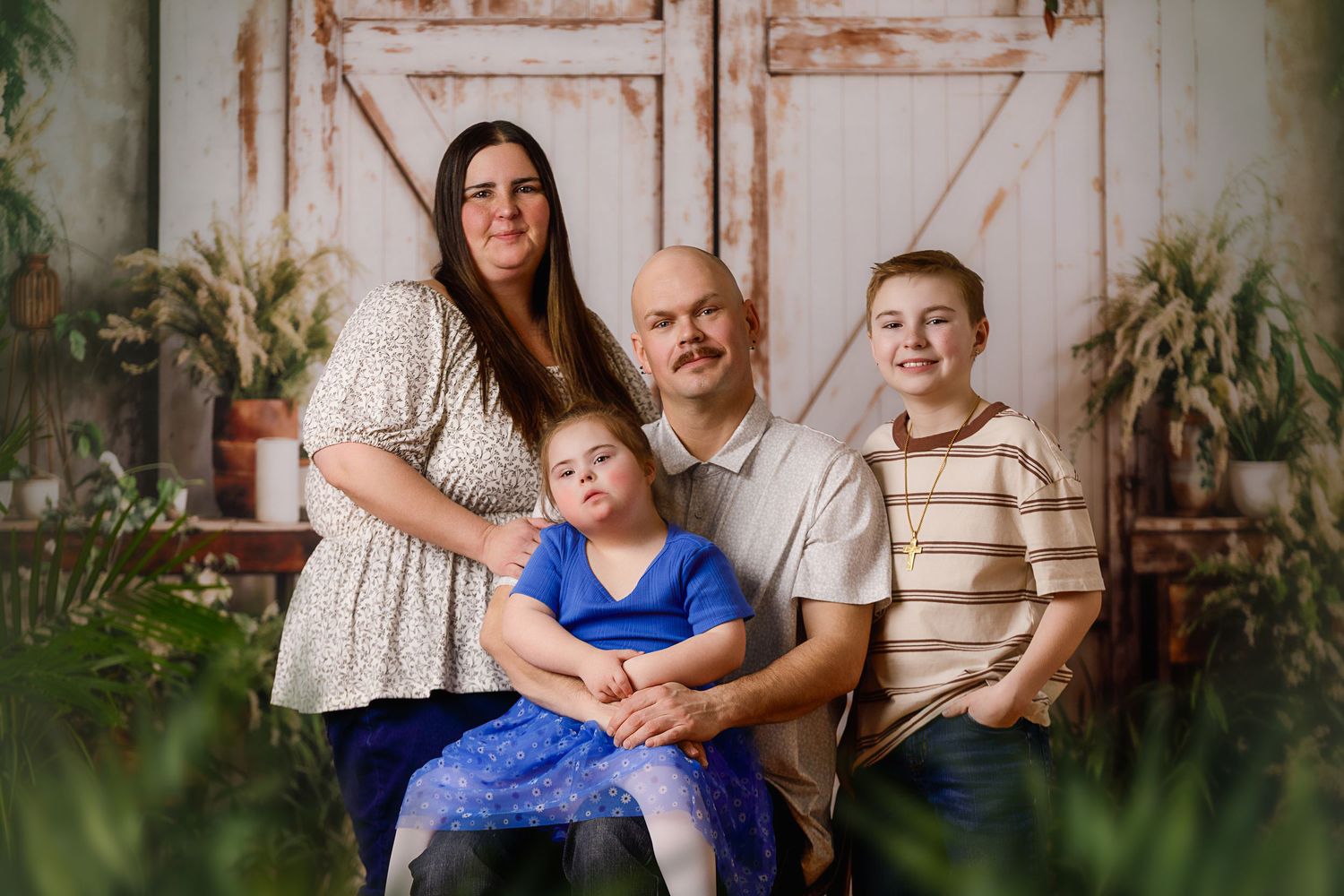 Family of four poses for a professional portrait in front of rustic barn doors surrounded by lush greenery.