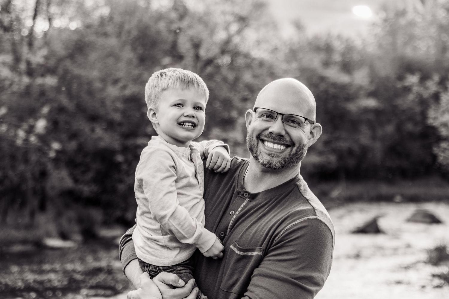Black and white photo of a bald father with glasses holding his laughing toddler son outdoors near trees.