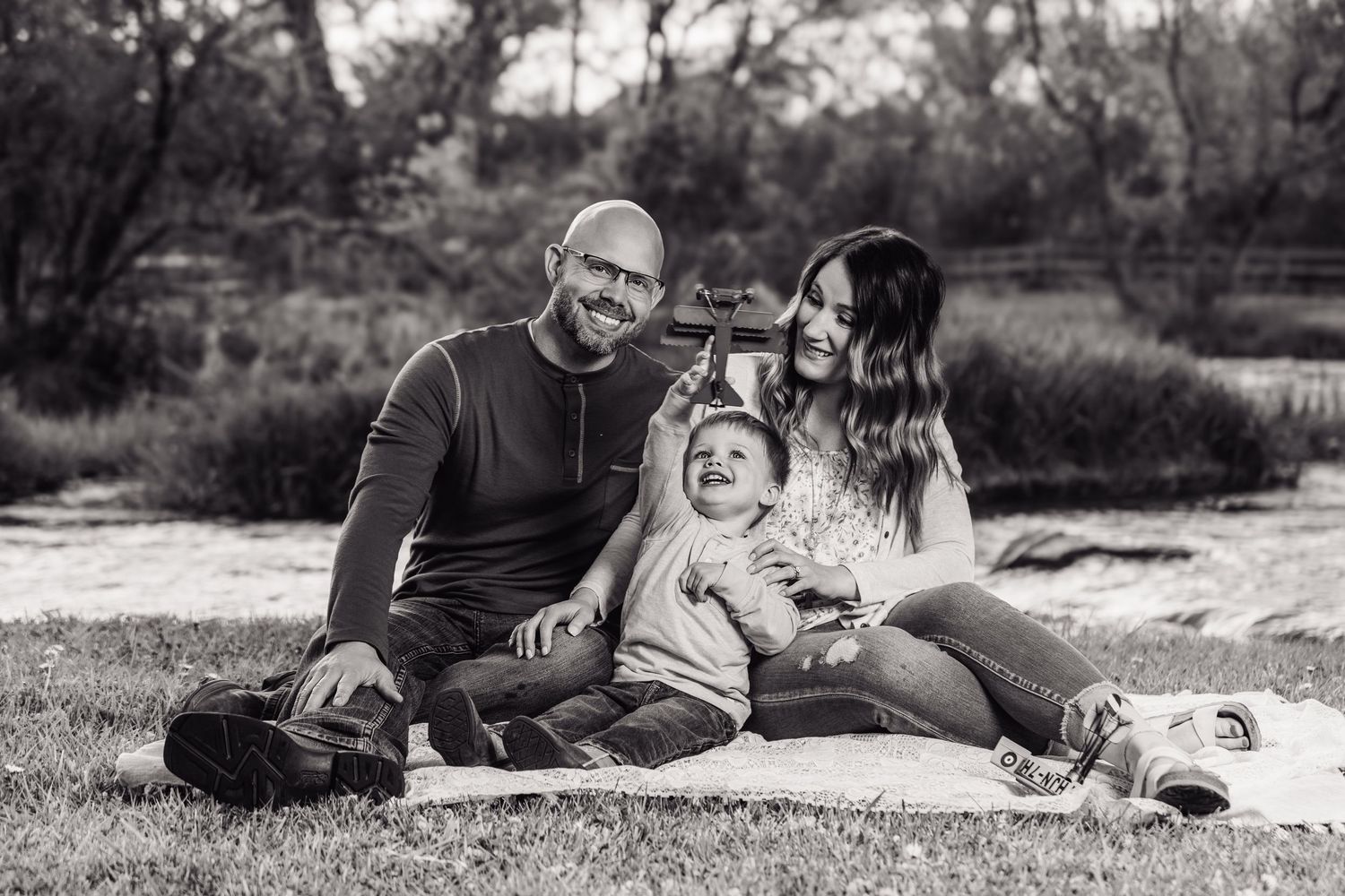Black and white photo of a happy family with a laughing baby sitting on a blanket outdoors near a river.