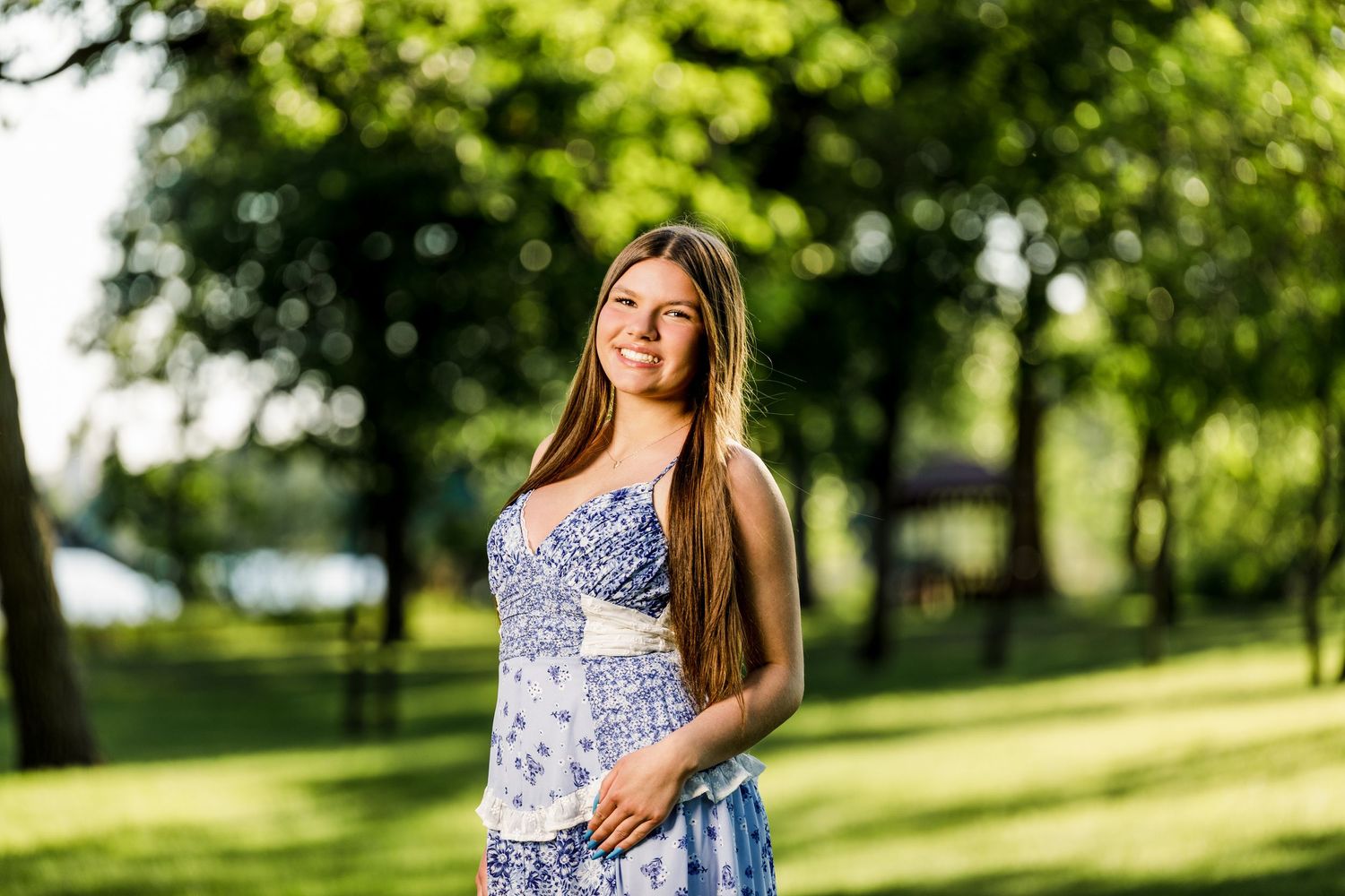Young woman in blue floral sundress smiling in lush green park during golden hour.