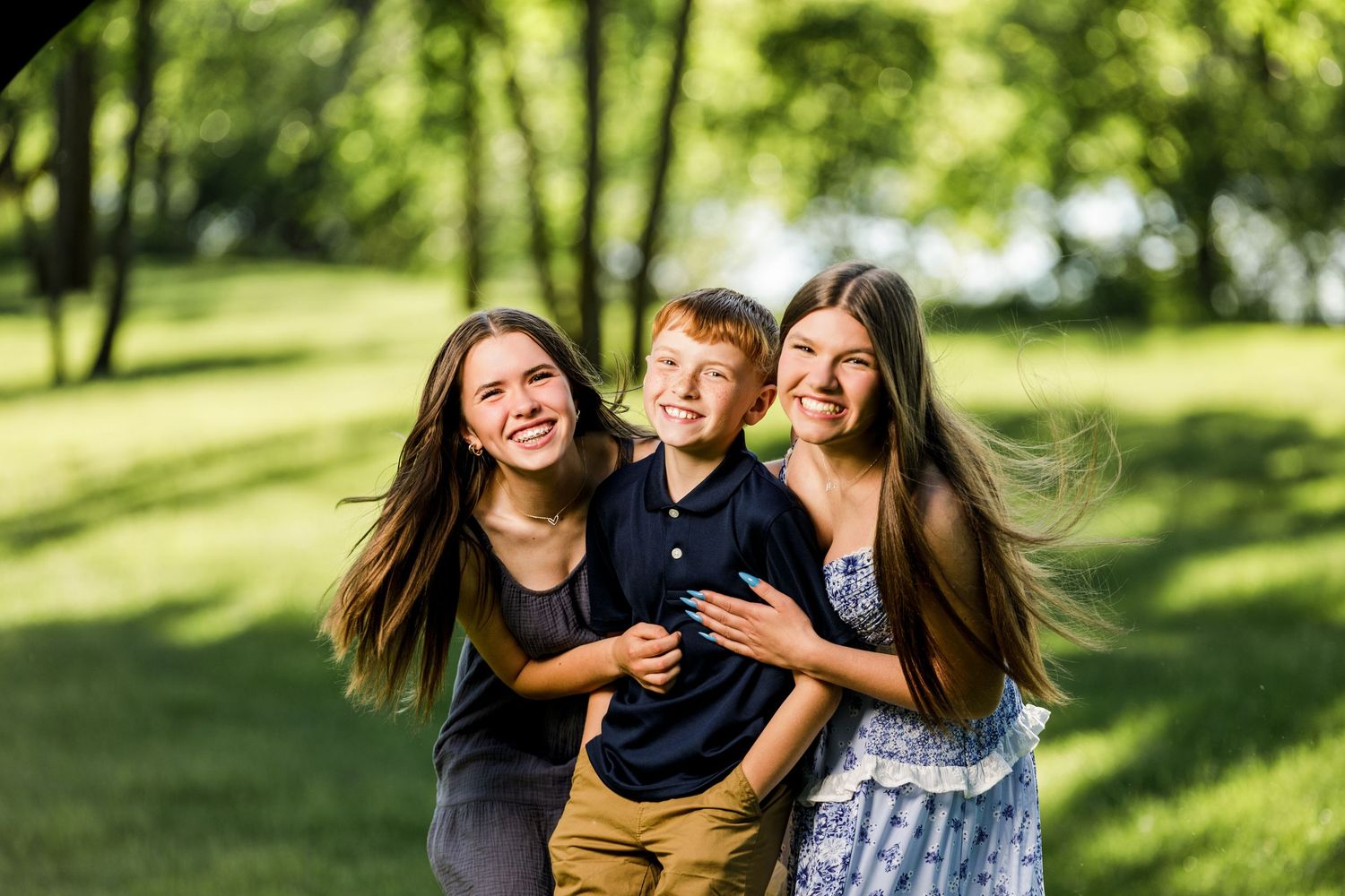 Two teenage girls hugging a young red-haired boy while laughing outdoors in a sunny green park.