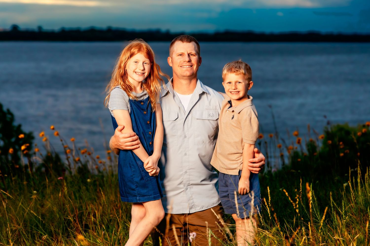 A family enjoys sunset together on a grassy hillside overlooking a tranquil lake during golden hour.