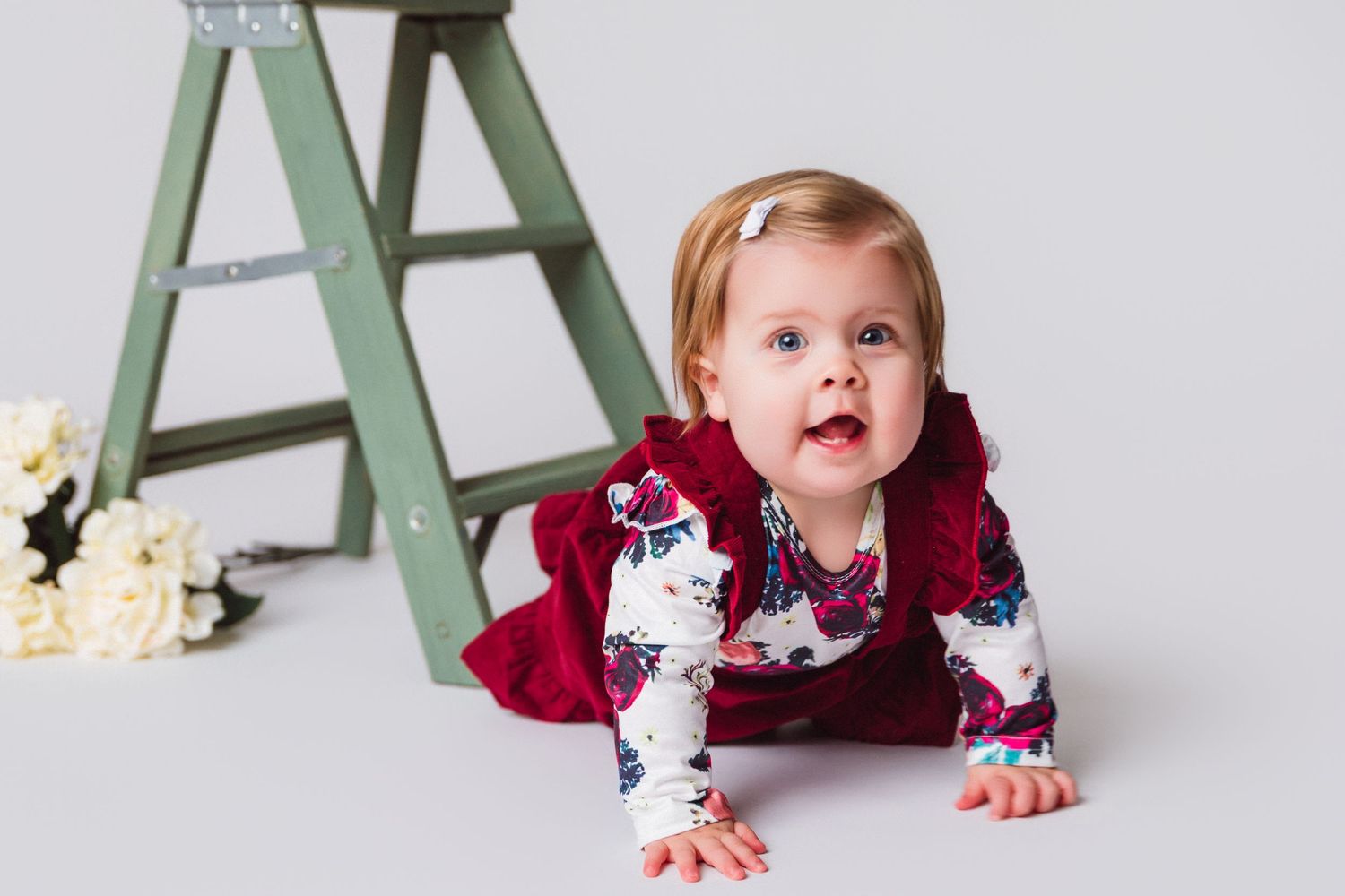 Baby in burgundy dress crawling and playing near mint green furniture.