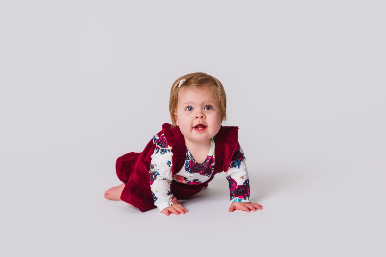 Baby in burgundy dress crawls and plays on light colored floor.