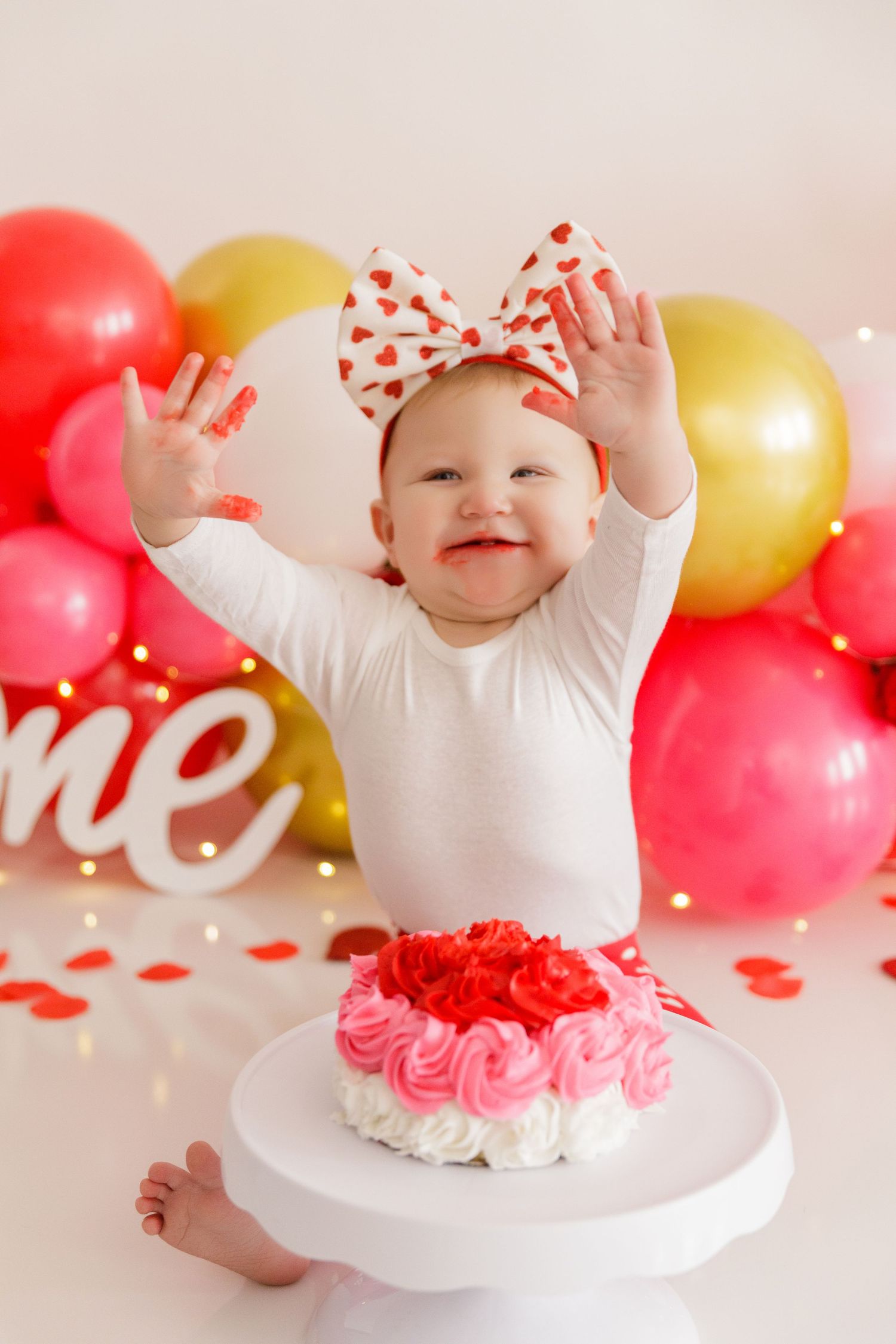 Baby in white outfit reaches up with joy near first birthday cake and balloon display.