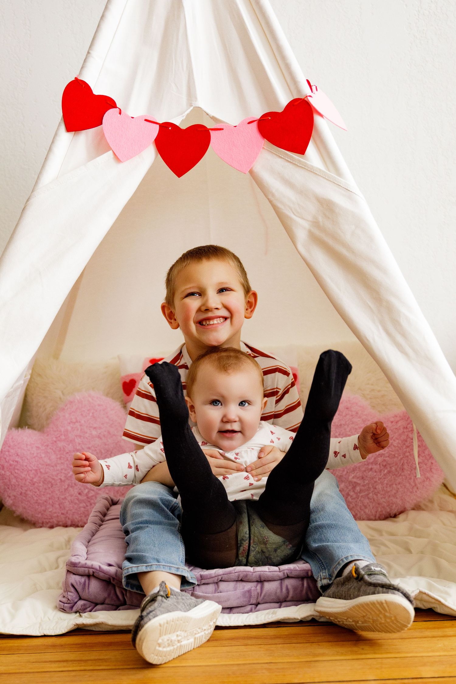 White teepee tent decorated with red heart garland and pink cushions on wooden floor.
