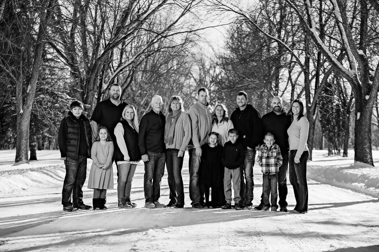 A large family group poses together on a snowy tree-lined path in black and white photography.