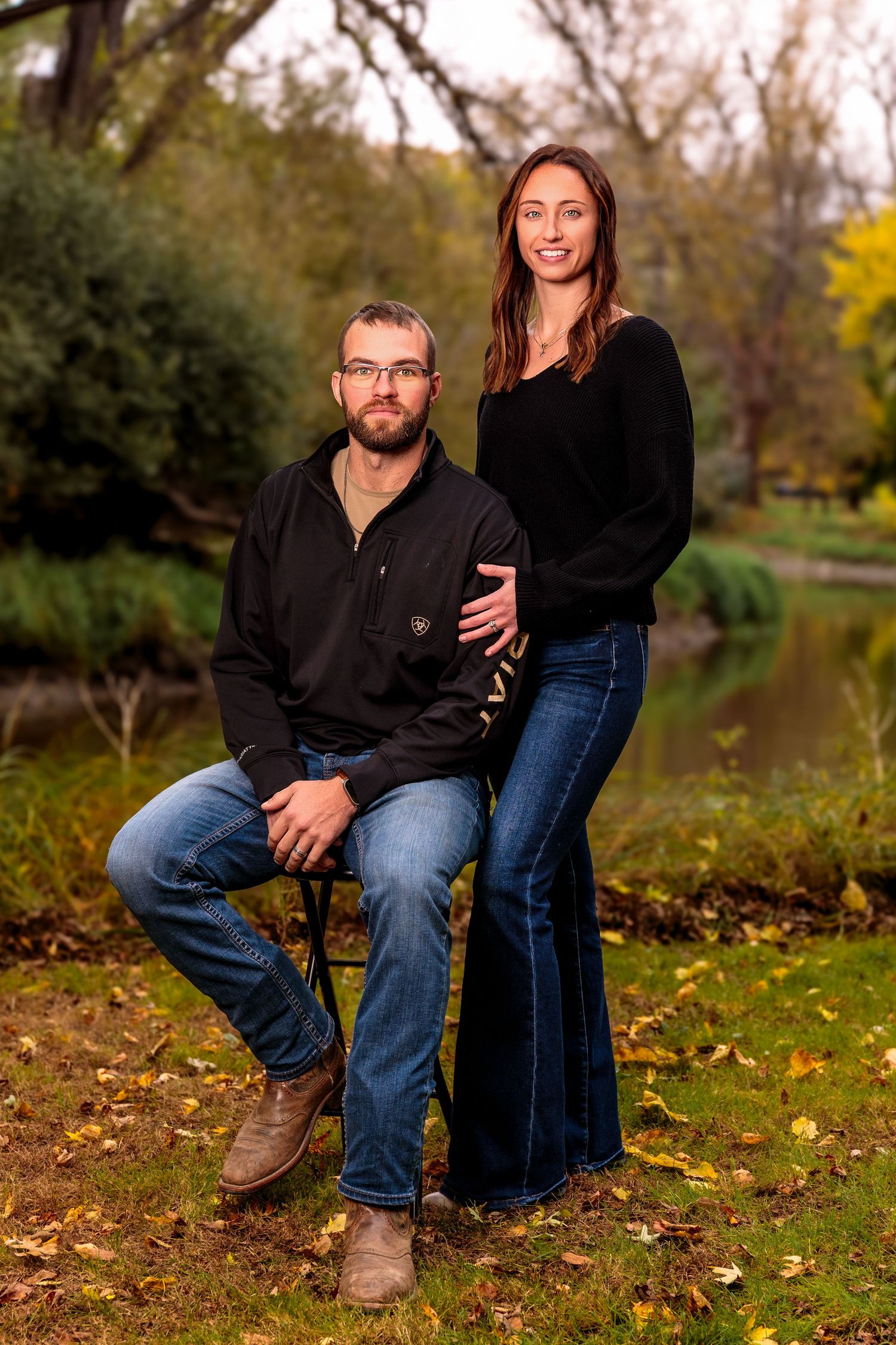 Couple poses on fallen autumn leaves near a pond during an outdoor photoshoot.