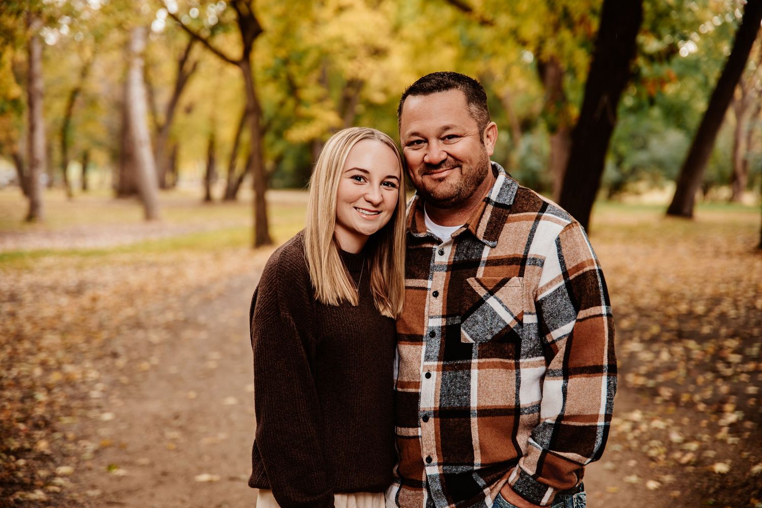 Couple poses together outdoors wearing casual fall attire among autumn trees.