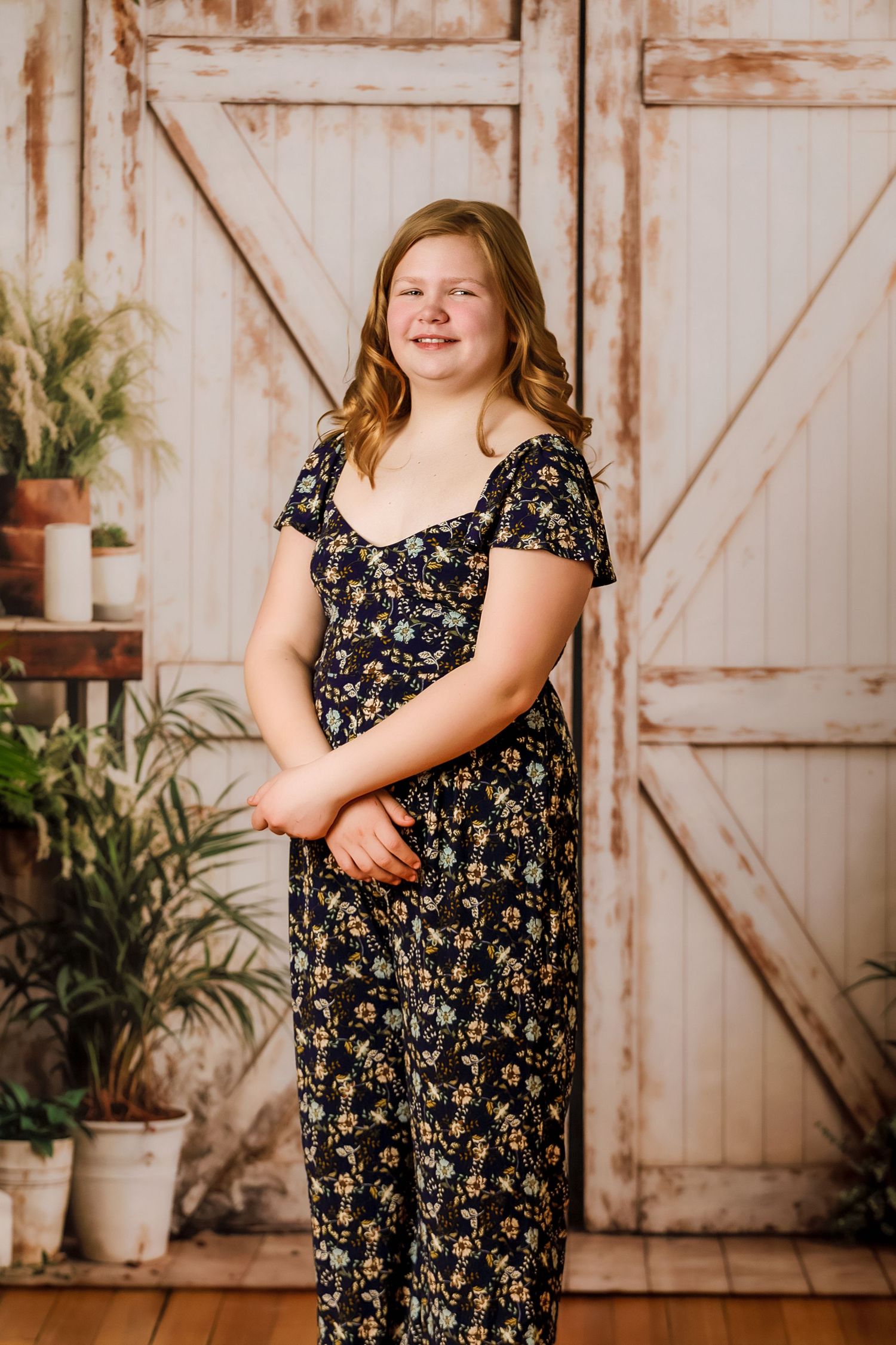 Smiling teenage girl in navy floral jumpsuit posing before rustic white barn door with potted plants.