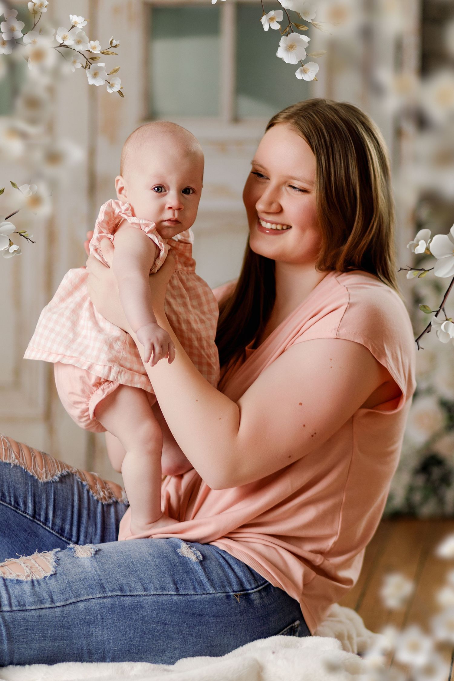 Smiling mother in pink shirt holds baby girl in pink dress among spring blossoms.